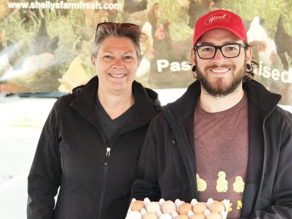 shelly_1 Two farmers stand together at a market, smiling and holding a carton of fresh eggs. The background features a banner promoting their farm, highlighting their focus on pasture-raised products.